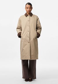 Μπεζ μακρύ παλτό με καφέ γιακά και μανίκια, διαθέτοντας κουμπιά μπροστά, πλευρικές τσέπες και χαλαρή σιλουέτα, σε συνδυασμό με καφέ παντελόνι.