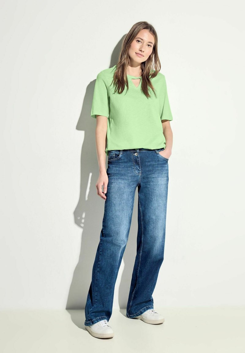 Grön kortärmad topp med ett framklipp, i kombination med blå vida jeans och vita sneakers. Enkel, avslappnad outfit på en neutral bakgrund.