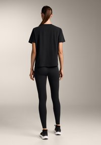 Žena stojící zády, oblečená v černém tričku, černých legínách a černých teniskách s bílou podešví na neutrálním pozadí.