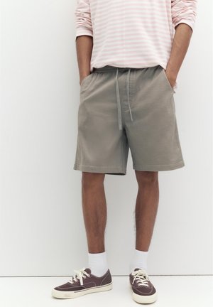 Person trägt beige Kordelzug-Shorts, weiße Socken, braune Sneakers und ein rosa-weiß gestreiftes langärmeliges Hemd mit den Händen in den Taschen.