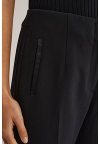 Svarta byxor i slätt tyg med framfickor med satinaccenter och ribbad midjeresår. Närbild på material och design.