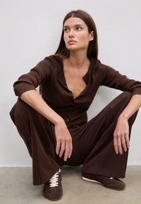 Ruskea uritettu pitkähihainen kardiganit, joissa napit, yhdistetty leveälahkeisiin ruskeisiin housuihin ja tummanruskeisiin lenkkareihin, joissa on valkoiset nauhat. Taustana on neutraali tausta.