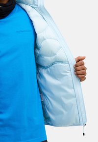 Lyseblå pufferjakke med quiltet tekstur, med ziplukning. Båret over en lys blå t-shirt med logodetalje.