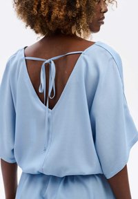 Jasnoniebieska bluzka z krótkimi, zwiewnymi rękawami, głębokim dekoltem z tyłu oraz ozdobnym wiązaniem na szyi, wykonana z miękkiego, gładkiego materiału.
