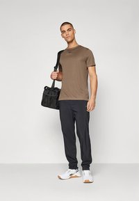 Brown athletic t-shirt, black athletic pants, and white sneakers. Model holds a black duffel bag. Neutral background.
