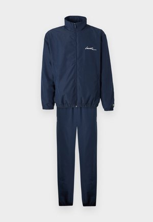 Navy blue tracksuit made of smooth fabric; features a zip-up jacket with high collar and elastic cuffs, and matching tapered pants with side zippers.