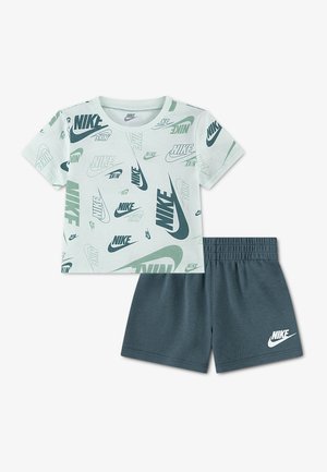 Camiseta blanca de manga corta de Nike con múltiples logotipos verdes y negros, combinada con pantalones cortos verdes oscuros de Nike con logo blanco en la pierna izquierda.