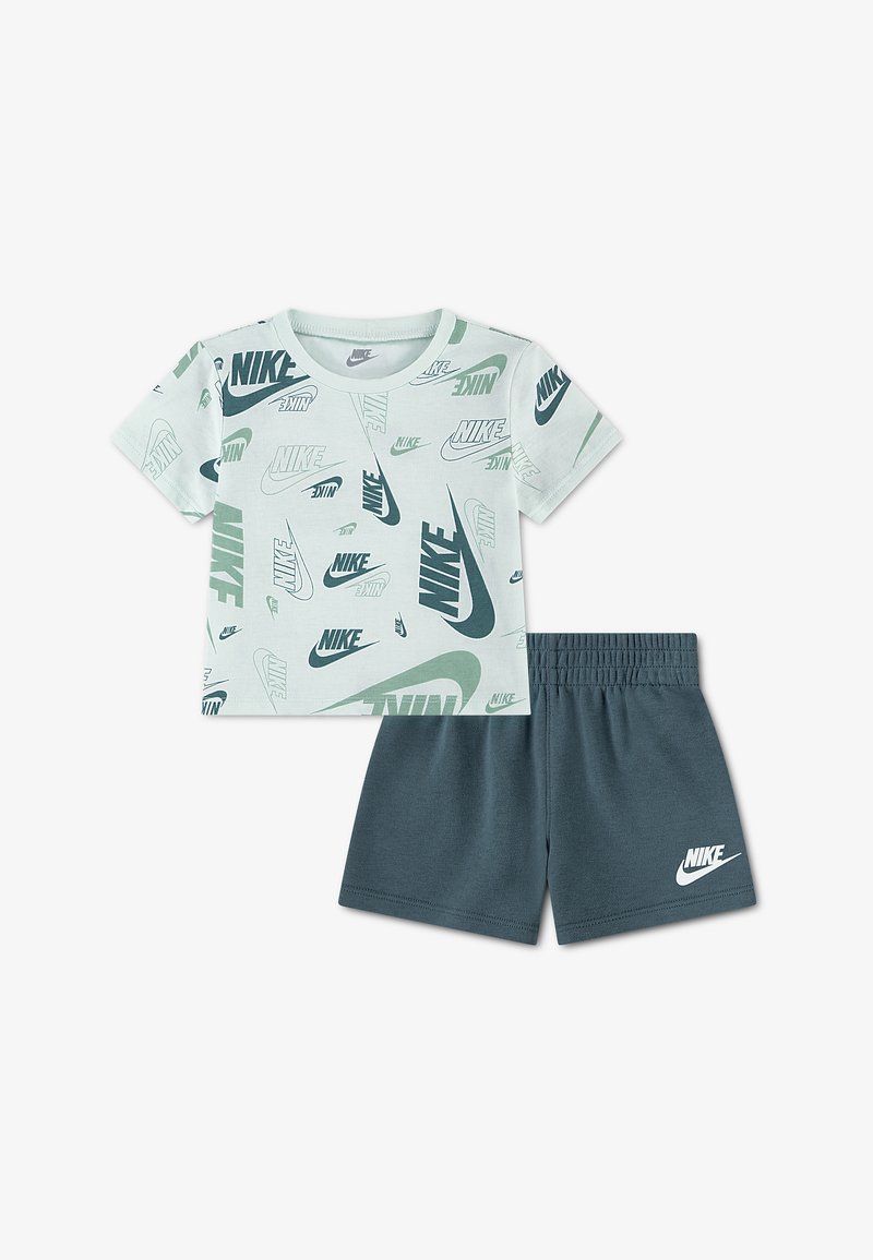 Λευκό κοντομάνικο t-shirt Nike με πολλαπλά πράσινα και μαύρα λογότυπα, συνδυασμένο με σκούρο πράσινο σορτς Nike με λευκό λογότυπο στο αριστερό πόδι.