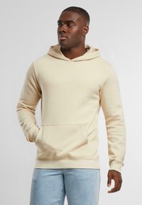 Felpa beige realizzata in tessuto morbido, con tasca a kanga, polsini a coste e cappuccio con cordino. Indossata su jeans azzurri chiari.