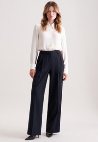 Donna con capelli mossi, che indossa una camicia bianca a bottoni, pantaloni neri a vita alta con gamba larga e scarpe col tacco nere a punta, in piedi davanti a uno sfondo neutro.