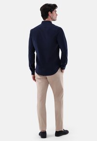 Camicia blu navy con colletto a punta, maniche lunghe e polsini; abbinata a pantaloni beige e scarpe nere, vista da dietro.