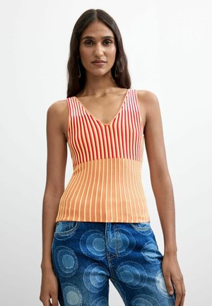 Vrouw draagt een mouwloze V-hals top met rode en oranje verticale strepen en blauwe jeans met cirkelpatronen, staand tegen een effen achtergrond.