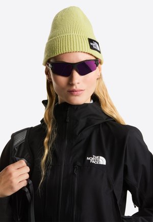 Zwarte jas met ritssluiting en getextureerde mouwen, lichtgroene gebreide muts en paars getinte zonnebril. Logo-label zichtbaar op de muts.