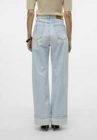 Šviesiai mėlyni plačios kojų džinsai su aukštu liemeniu, turintys briaunotą apdailą ir dvi užpakalines kišenes, derinami su smėlio spalvos palaidine ir aukštakulniais batais.