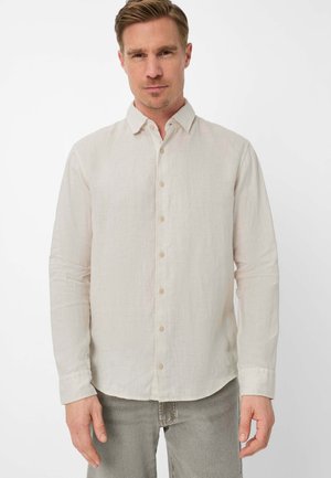 Mann trägt ein hellbeiges, langärmliges Hemd mit Knöpfen und graue Hose, steht vor einem schlichten weißen Hintergrund.