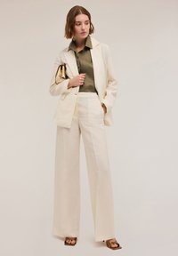 Blazer oversize beige chiaro e pantaloni a gamba larga abbinati, indossati con una camicia button-up verde oliva scuro. Tiene una clutch dorata.
