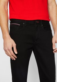Jeans de denim negro con corte slim, que presentan un acento rojo en el bolsillo, diseño de cinco bolsillos y detalles de costura en contraste.