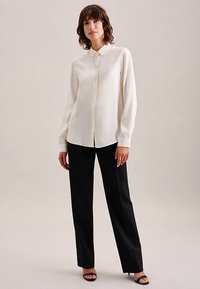 Licht crèmekleurig blouse met lange mouwen, een knoopsluiting aan de voorkant en een klassieke kraag, gecombineerd met zwarte, op maat gemaakte broek en zwarte sandalen.