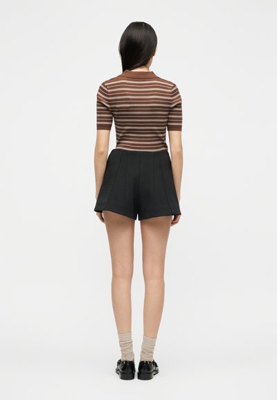 Γυναίκα με μακριά σκούρα μαλλιά, φοράει καφέ ριγέ κοντομάνικο πουκάμισο, μαύρο σορτς ψηλόμεσο, μπεζ κάλτσες και μαύρα παπούτσια, στέκεται γυρισμένη προς τα πίσω.