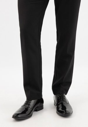 Schwarze lackierte Lederschuhe, getragen zu schwarzen formellen Hosen, vor weißem Studiohintergrund.