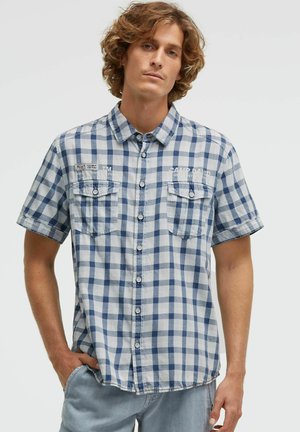 Junger Mann mit lockigem Haar, der ein blau-weiß kariertes Kurzarmhemd und hellgraue Hose trägt, steht mit einer Hand in der Tasche.