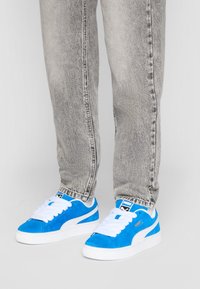 Baskets en suède bleu avec des accents et des lacets blancs, présentant une finition texturée. Portées avec un jean tapered gris clair.