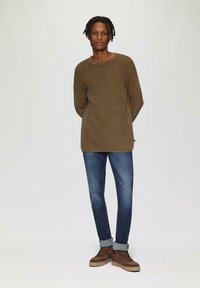 Pull marron texturé à la coupe ample, porté sur un jean en denim bleu. Des chaussures montantes marron et un jean retroussé complètent la tenue.