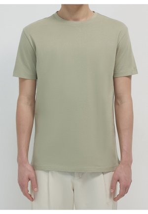 Hombre con camiseta de manga corta color verde oliva claro lisa y pantalones color blanco roto, de pie frente a un fondo liso.