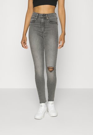 Personne portant un jean skinny gris taille haute avec une déchirure au niveau d'un genou, des baskets blanches et un top court noir sur un fond uni.