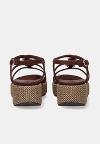 Sandalias de tiras de ante marrón con hebillas doradas y plataformas trenzadas, vistas desde atrás sobre un fondo blanco.