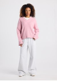 Maglione rosa a coste con scollo a V, abbinato a pantaloni della tuta larghi di colore grigio chiaro. La modella è in piedi contro uno sfondo semplice.