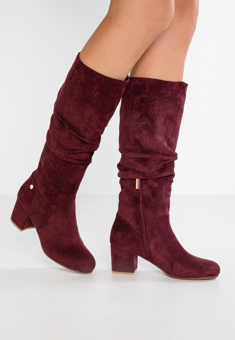 Bottes en suede bourgogne montantes avec un design flou, talon bloc épais, fermeture éclair sur le côté et détails en or. Texture douce et finition lisse.