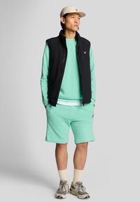 Chaleco acolchado negro, sudadera verde menta, pantalones cortos verdes menta, calcetines crema y zapatillas. Gorra con logo y chaleco frontal con cremallera.