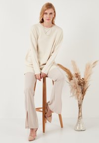 Pull en côte crème, pantalon beige clair et talons beiges. Teintes neutres chaudes. Sur fond minimaliste avec des accents floraux séchés.