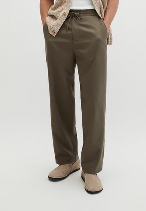 Person trägt olivgrüne Kordelhose, beige Slipper, weißes geripptes Oberteil und eine beige strukturierte offene Weste, steht mit den Händen in den Taschen.