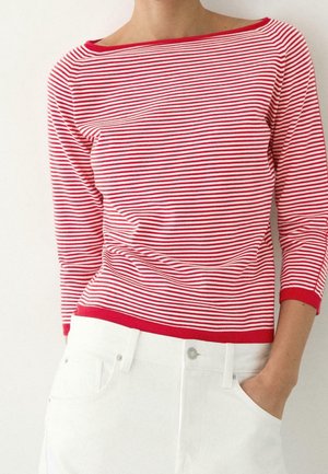 Personne portant une chemise à manches longues rayée rouge et blanche avec un pantalon blanc, une main dans la poche, sur un fond blanc uni.