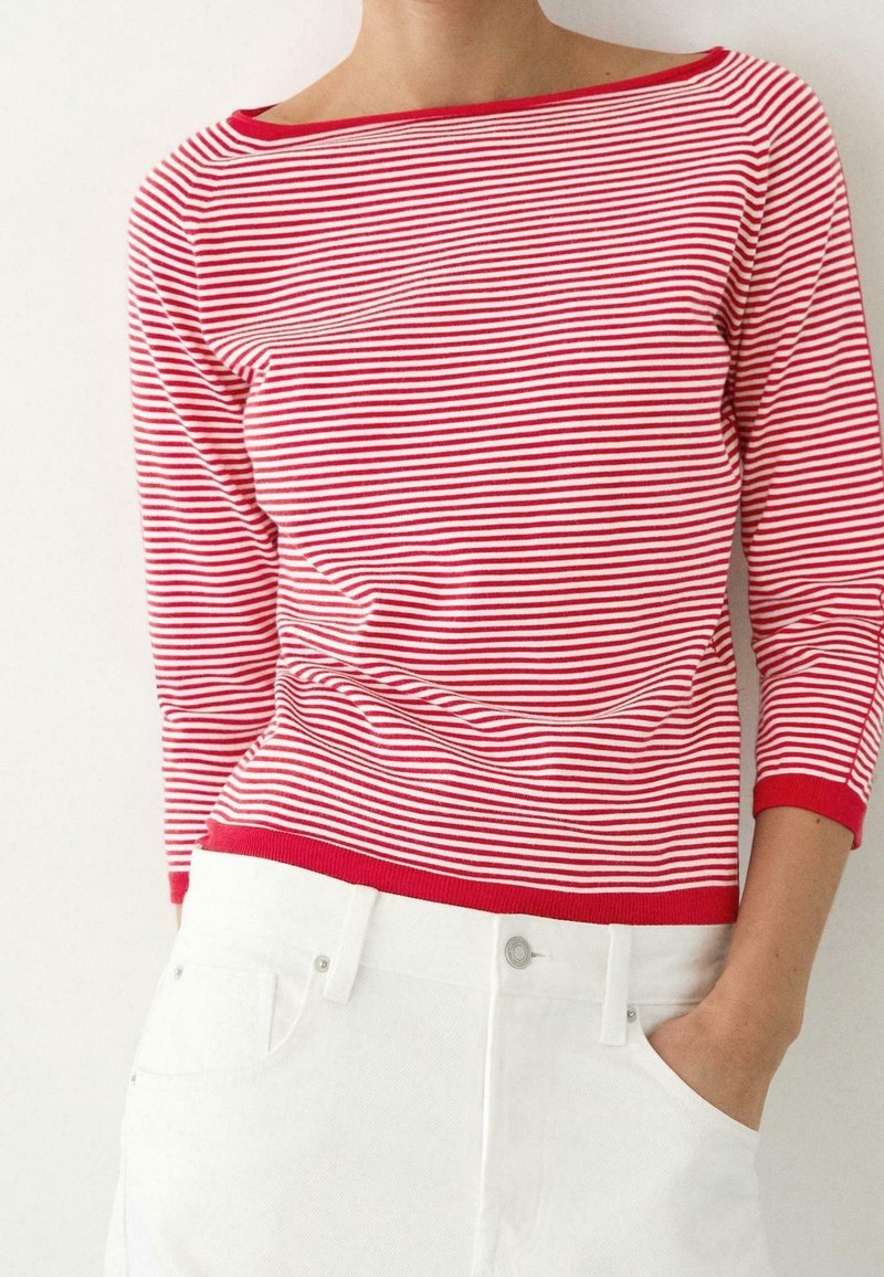 Personne portant une chemise à manches longues rayée rouge et blanche avec un pantalon blanc, une main dans la poche, sur un fond blanc uni.