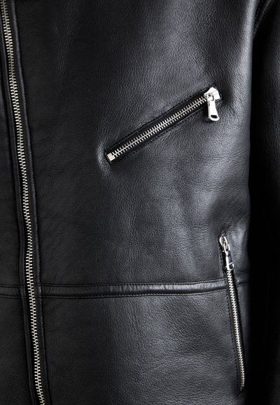 Svart läderjacka med flera dragkedjefickor, slät yta och en strömlinjeformad design. Silverfärgade detaljer accentuerar dragkedjorna.