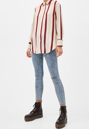 Kobieta ubrana w kremową koszulę z długim rękawem w pionowe czerwone paski, jasnoniebieskie podarte obcisłe dżinsy oraz czarne sznurowane buty na platformie.