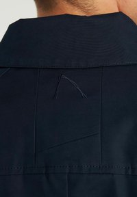 Närbild av bakkragen på en mörkt marinblå jacka bärs av en person med kort hår, med en liten broderad abstrakt logo.