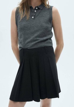 Femme portant un polo sans manches gris foncé rentré dans une jupe plissée noire taille haute, debout, les mains derrière le dos.