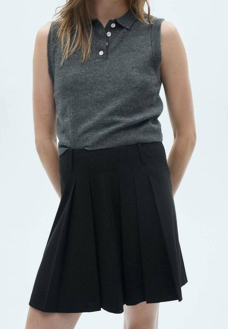 Femme portant un polo sans manches gris foncé rentré dans une jupe plissée noire taille haute, debout, les mains derrière le dos.