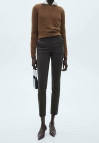 Pull marron à col rond, associé à un pantalon gris ajusté. Le mannequin tient un petit sac à main noir, rehaussé par des talons aiguille bourgogne.