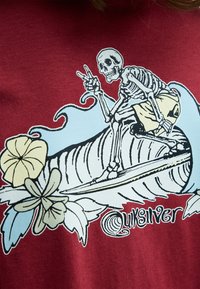 Skelet surfer på en bølge med fredstegn, omgivet af tropiske blomster, på et rødt stof med "Quiksilver" logo.