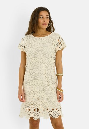 Femme portant une robe beige en crochet à manches courtes avec des motifs floraux, regardant sur le côté, avec des bracelets à son poignet droit.