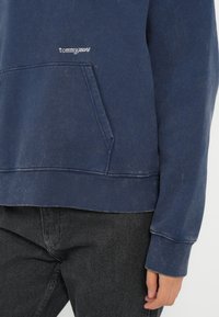 Mørkeblå bomullshoodie med kengurulomme, ribbede mansjetter og linning. Har hvit "tommy jeans" logo broderi på brystet.