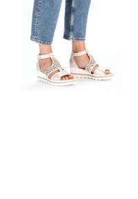 Beige wedge sandals with cutout design and ankle strap, featuring a white sole and textured finish. Paired with cropped blue jeans.