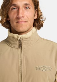 Veste zippée beige avec un col haut, doublure en polaire texturée et logo brodé sur la poitrine. Elle possède un tissu lisse et léger.