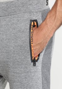 Grijze sweatpants met een zijzak voorzien van een oranje ritssluiting en zwarte details. De stof heeft een zachte textuur en een relaxte pasvorm.