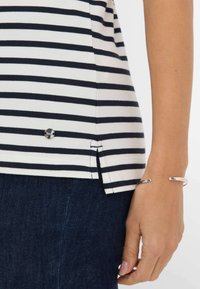 Top a righe blu navy e bianche con spacco laterale e dettaglio argentato, abbinato a jeans scuri. Tessuto morbido e design casual.